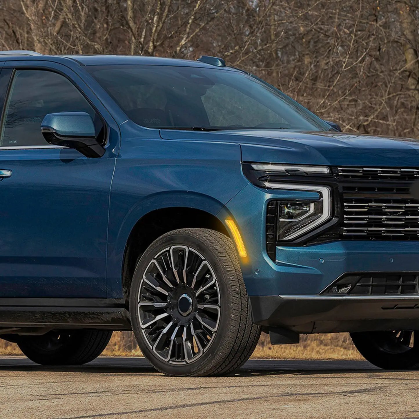 Close-up view of a blue SUV parked outdoors, highlighting the front wheel, modern alloy rim, illuminated amber side marker light, and sleek headlight design with black grille.