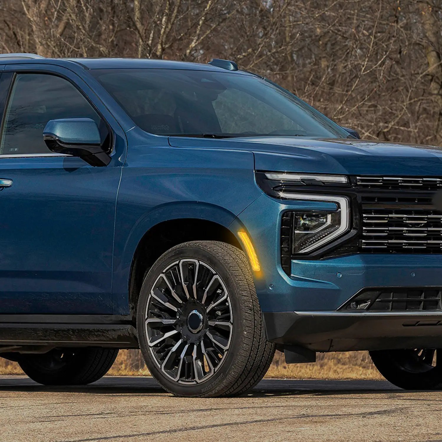 Close-up view of a blue SUV parked outdoors, highlighting the front wheel, modern alloy rim, illuminated amber side marker light, and sleek headlight design with black grille.